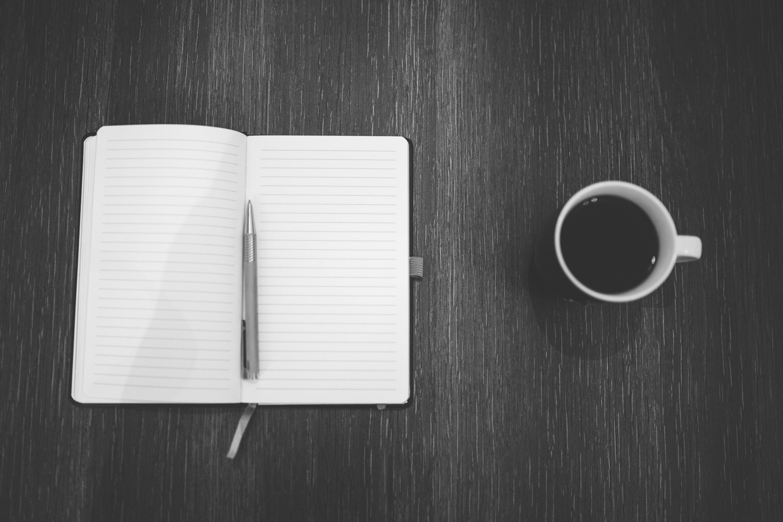 A black and white image. To the left, a notebook open to blank pages with a pen laying in its spine and a coffee cup to the right.